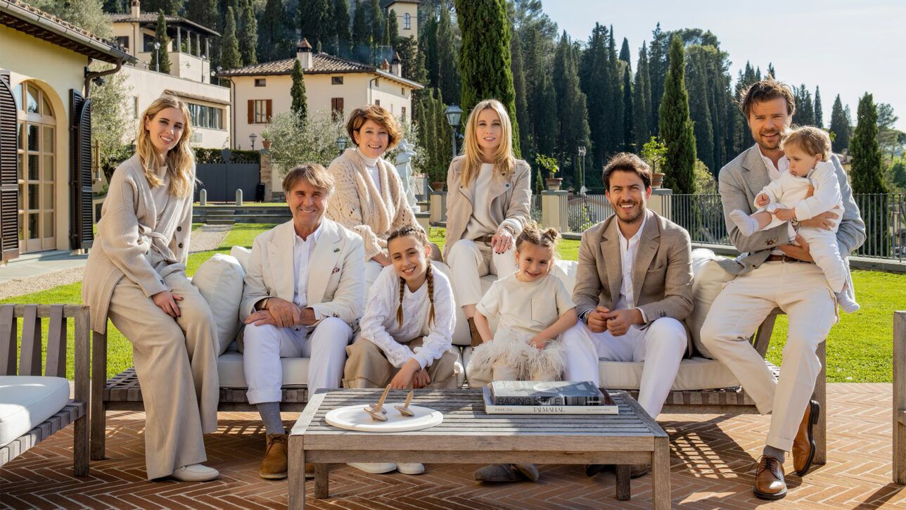 Cream and tan linen tailoring, knitwear and trousers worn by a family group on a terrace, a styled lifestyle scene