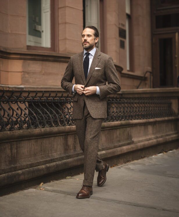 Dark brown donegal tweed suit with blue shirt, navy tie and brown brogues, worn by a model on a city street