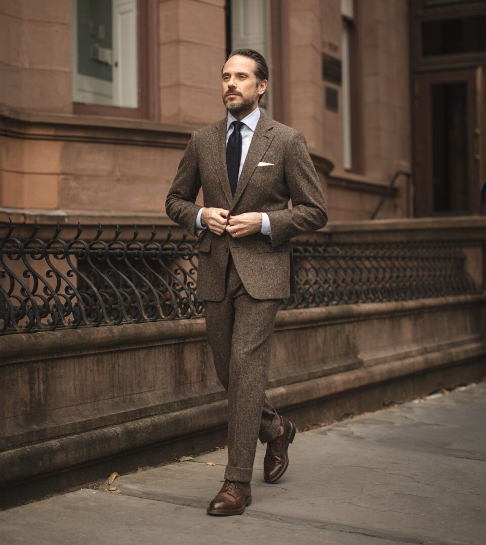 Dark brown donegal tweed suit with blue shirt, navy tie and brown brogues, worn by a model on a city street