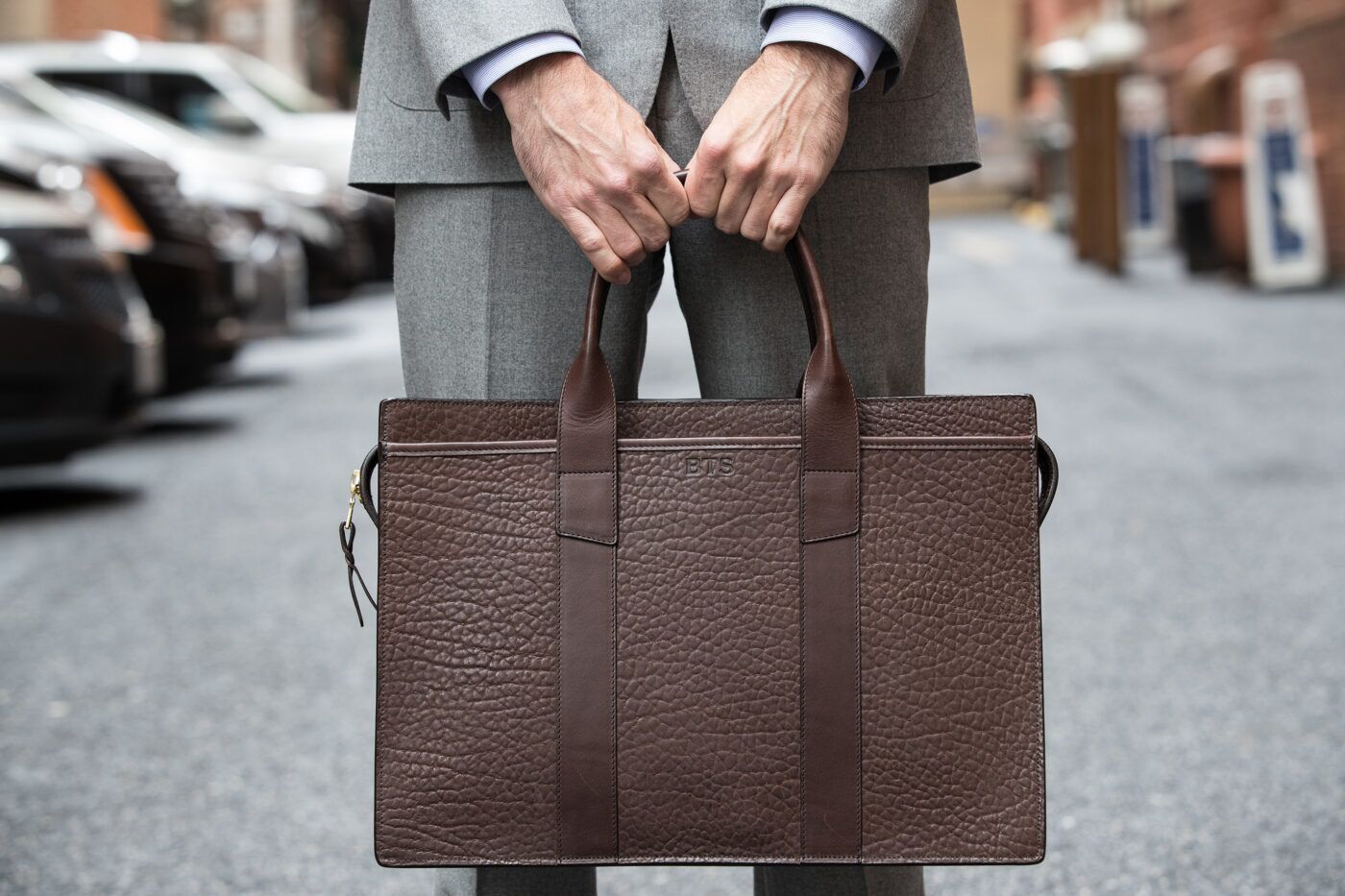 Dark brown shrunken grain leather zip-top briefcase with monogram, carried by a model in a grey suit