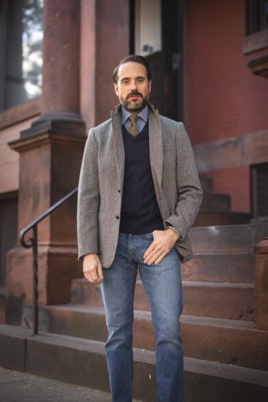 Grey herringbone tweed sport coat with navy V-neck sweater, chambray shirt, olive tie and jeans, worn by a model