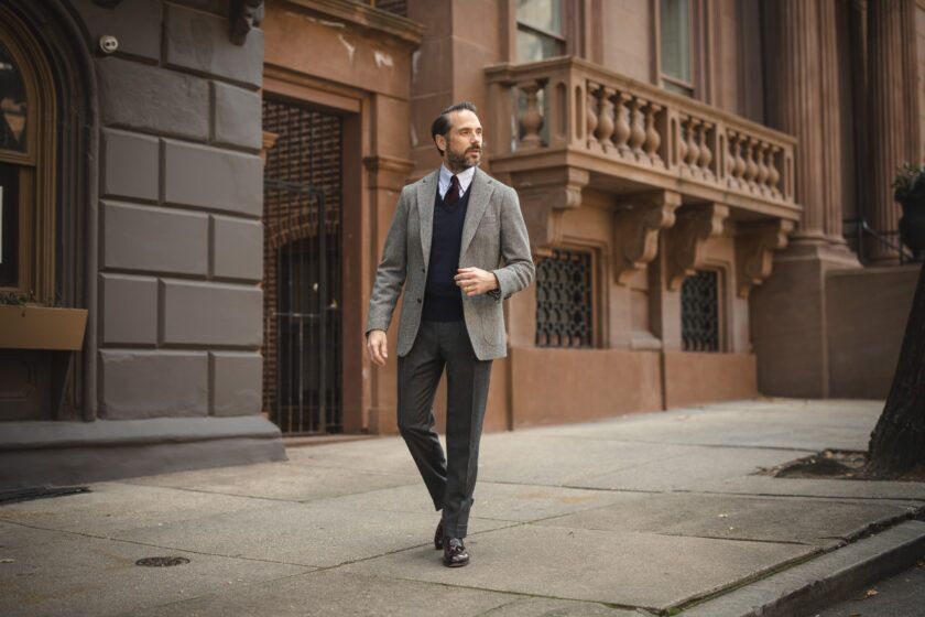 Grey tweed sport coat with navy V-neck sweater, burgundy tie and charcoal flannel trousers, worn by a model on a city street