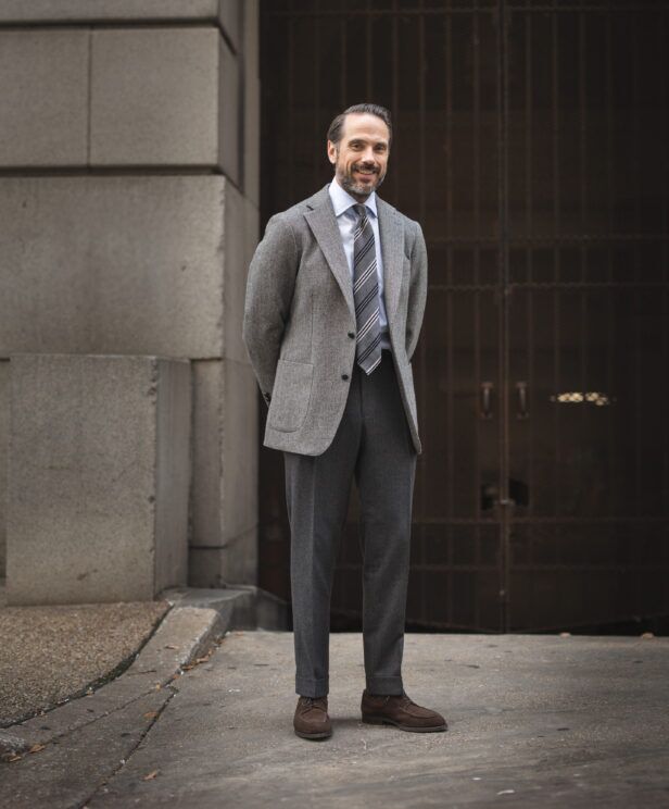 Grey tweed sport coat with striped tie, blue shirt, charcoal flannel trousers and brown suede shoes, worn by a model