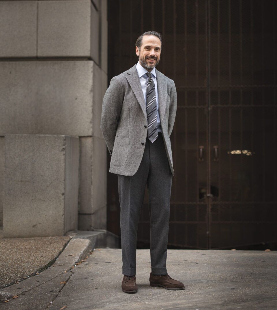 Grey tweed sport coat with striped tie, blue shirt, charcoal flannel trousers and brown suede shoes, worn by a model