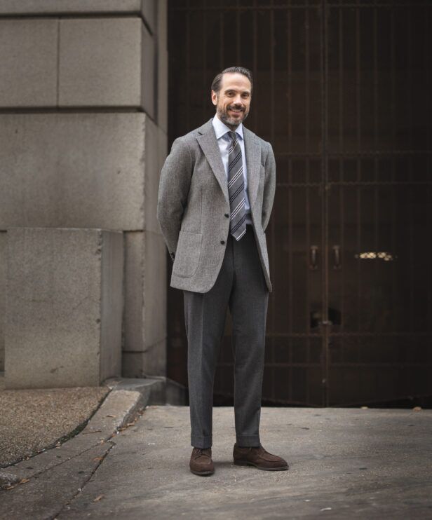 Grey tweed sport coat with striped tie, blue shirt, charcoal flannel trousers and brown suede shoes, worn by a model