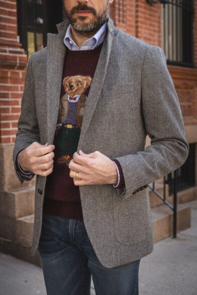 Grey herringbone tweed sport coat with burgundy bear-motif sweater, blue shirt and jeans, worn by a model on a city street