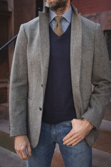 Grey herringbone tweed sport coat with navy V-neck sweater, chambray shirt, brown knit tie and jeans, worn by a model
