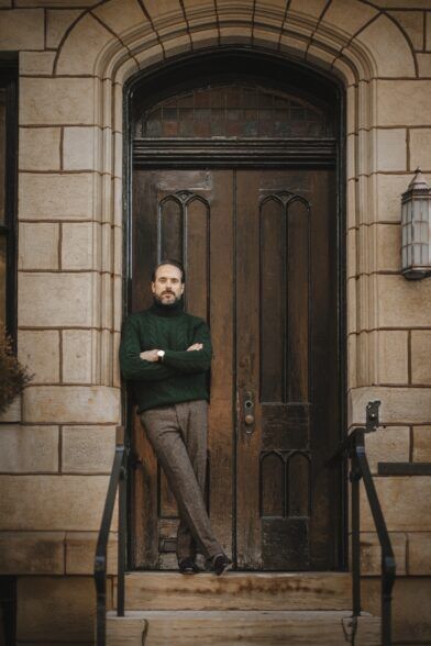 Green cable knit turtleneck with brown tweed trousers and suede loafers, worn by a model in a stone doorway