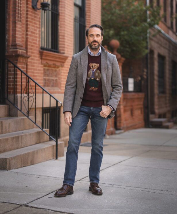 Gray tweed sport coat with burgundy teddy bear sweater, blue jeans and brown brogues, worn by a model on a city sidewalk