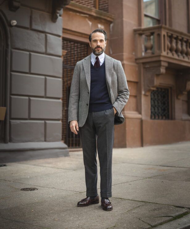 Grey tweed sport coat with navy V-neck sweater, burgundy tie and charcoal flannel trousers, worn by a model on a city street