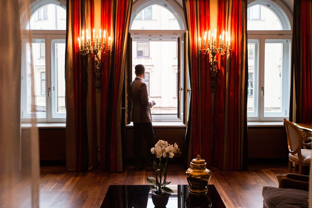 Brown sport coat with charcoal trousers, worn by a man at an arched window in a hotel suite, styled lifestyle scene