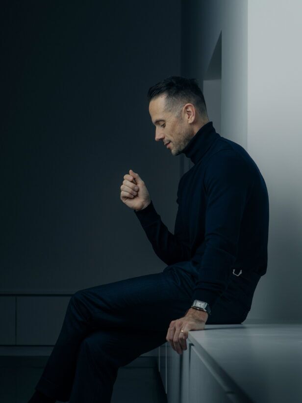 Navy fine-knit turtleneck with dark navy trousers and a rectangular steel watch, worn by a seated model