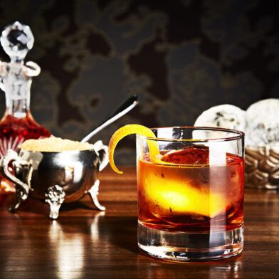 Old fashioned cocktail with orange twist and large ice cube beside a silver sugar bowl and decanter, styled bar scene