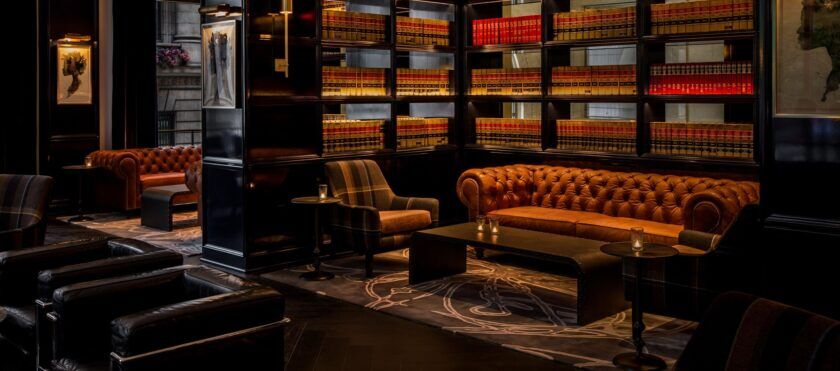 Library-style cocktail bar lounge with tan leather chesterfield sofas and lit bookshelves, a lifestyle scene