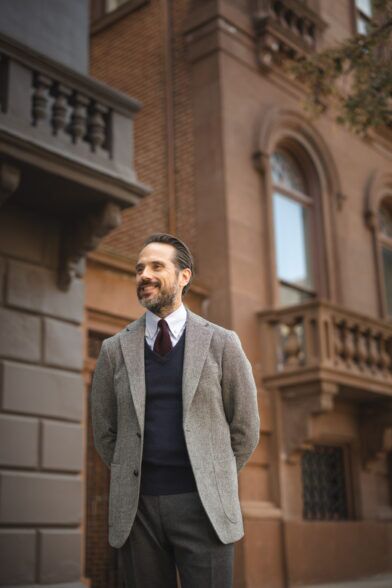 Grey tweed sport coat with navy V-neck sweater, white shirt and burgundy tie, worn by a model on a city street