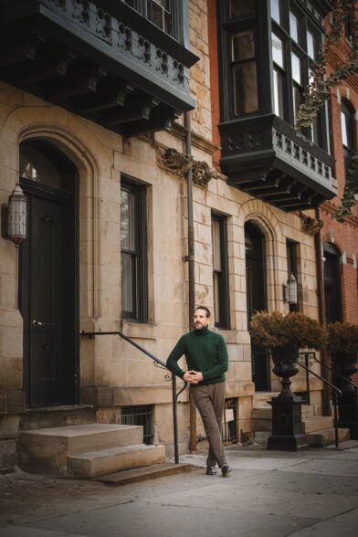 Green cable knit turtleneck sweater with brown tweed trousers and loafers, worn by a model on a brownstone street