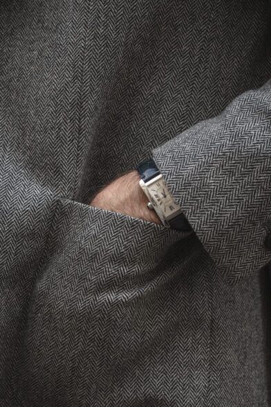 Grey herringbone tweed coat with patch pocket and rectangular watch on navy leather strap, a close-up detail