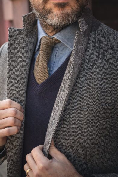 Grey herringbone tweed coat with olive knit tie, chambray shirt and navy V-neck sweater, a close-up on a model