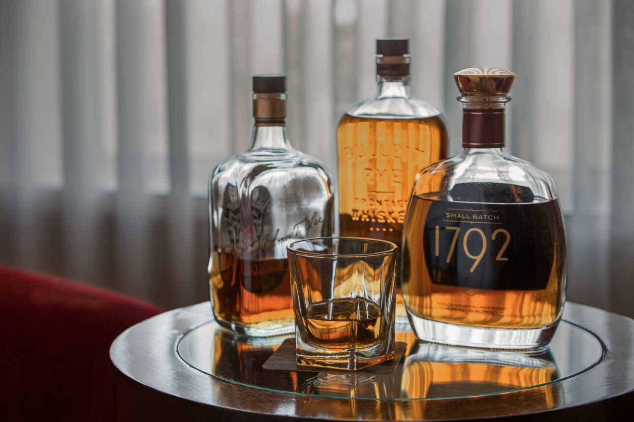Bourbon bottles and a poured rocks glass on a mirrored tray beside a red velvet chair, a styled lifestyle scene
