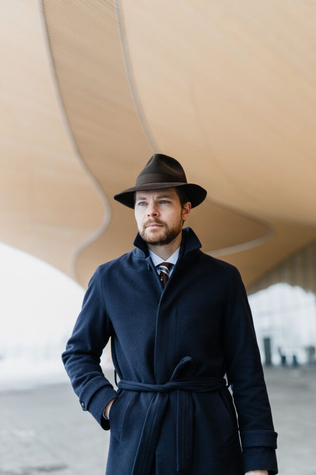 Navy wool belted overcoat with burgundy striped tie and brown felt fedora, worn by a model outdoors