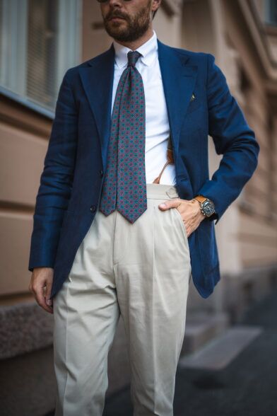 Navy sport coat with green and pink medallion silk tie, white shirt and cream pleated trousers, worn by a model