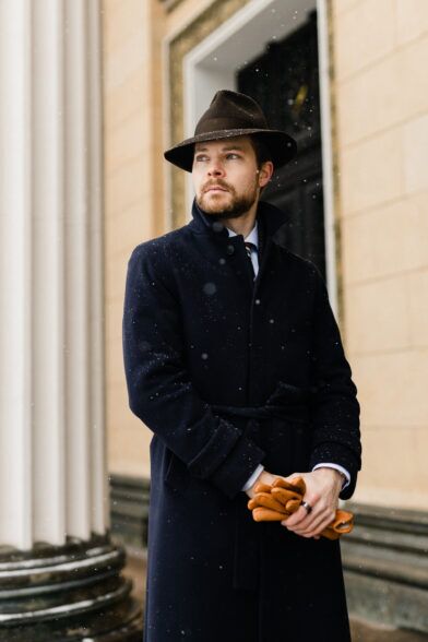 Navy wool belted overcoat with brown felt fedora and tan leather gloves, worn by a model in falling snow