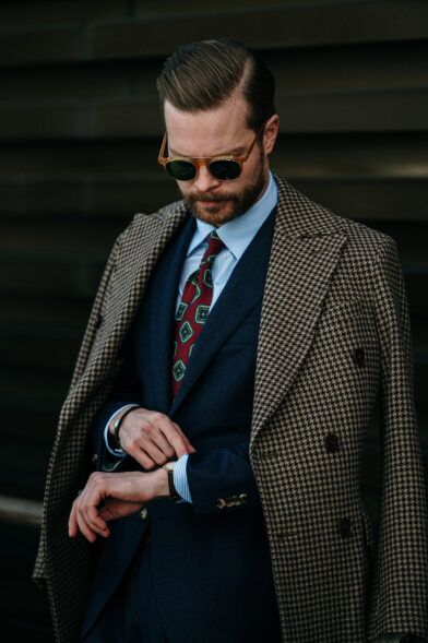 Brown houndstooth wool overcoat draped over a navy suit, striped shirt and burgundy medallion tie, worn by a model