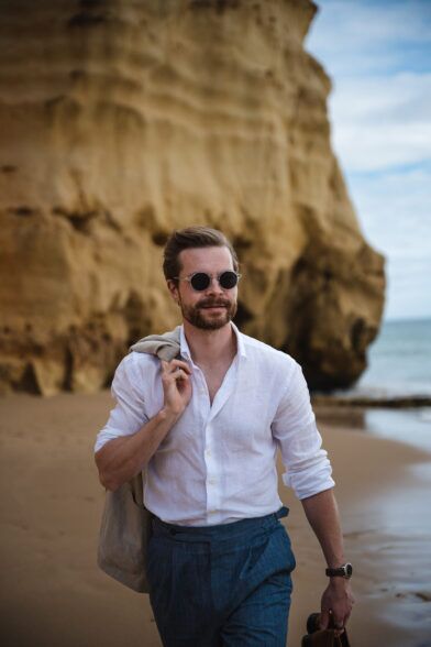 White linen shirt with rolled sleeves and blue pleated linen trousers, with a tan jacket over the shoulder, on a beach