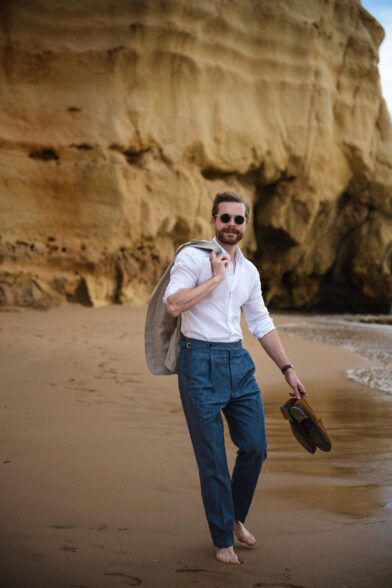 Blue pleated linen trousers with white linen shirt and tan jacket over the shoulder, worn barefoot on a beach