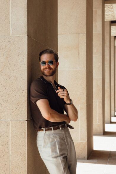 Brown knit polo shirt with contrast tipping and stone pleated linen trousers, in a sunlit colonnade