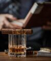 Banded cigar resting on a cut-glass tumbler of whiskey with a gold lighter and playing cards, a styled lifestyle scene