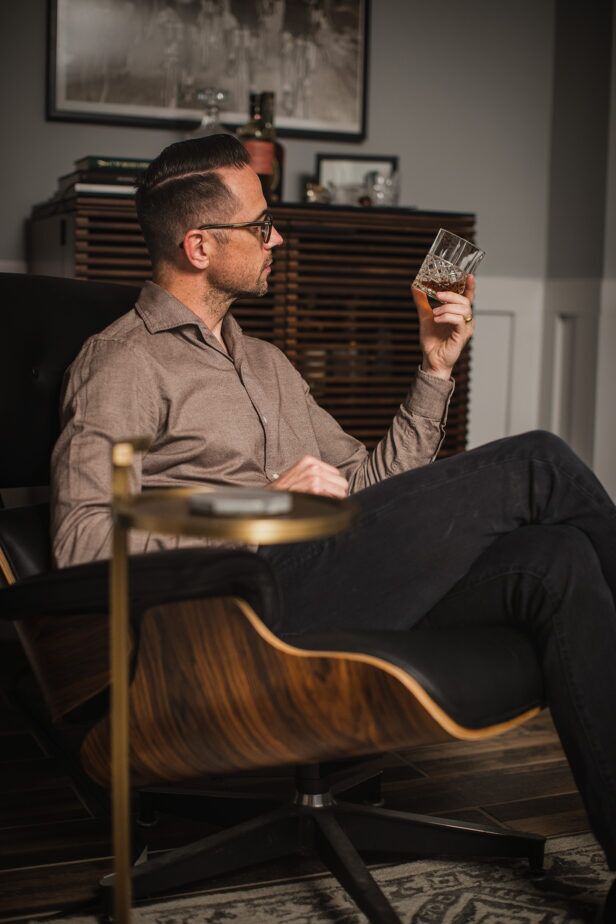 Taupe brushed cotton spread collar shirt with dark denim, worn by a seated model holding a whiskey glass