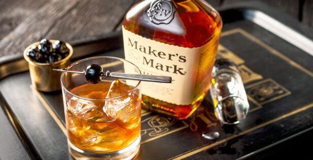 Bourbon cocktail on ice with a cherry garnish beside a Maker's Mark bottle, a styled lifestyle scene