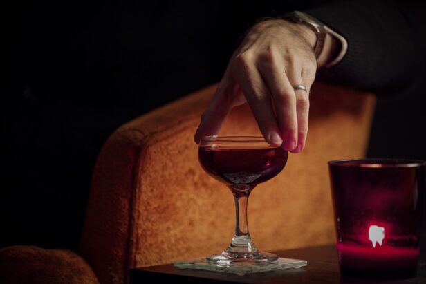 Steel watch and plain band ring worn with a dark sleeve, hand reaching for a Manhattan cocktail in a candlelit bar scene