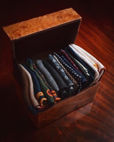 Folded pocket squares in navy, green, grey and white patterns stored in an open wooden box, styled scene