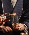 Navy quilted smoking jacket with cream piping, worn while bourbon is poured into a tasting glass, lifestyle scene