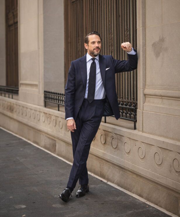 Navy flannel two-piece suit with light blue shirt, navy grenadine tie and black oxfords, worn by a model on a city street