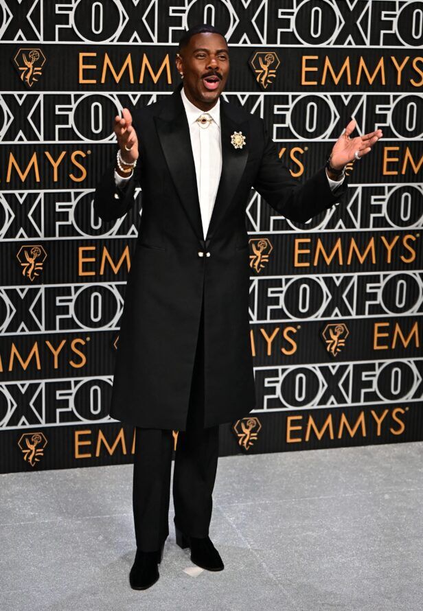 Black longline tuxedo coat with satin peak lapels, white shirt and gold brooch, worn on a red carpet
