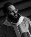 Shearling overcoat with cable knit turtleneck and knotted scarf, worn by a bearded man, black and white street scene