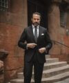 Black flannel suit with white shirt, grey tie and white pocket square, worn by a model on a city street