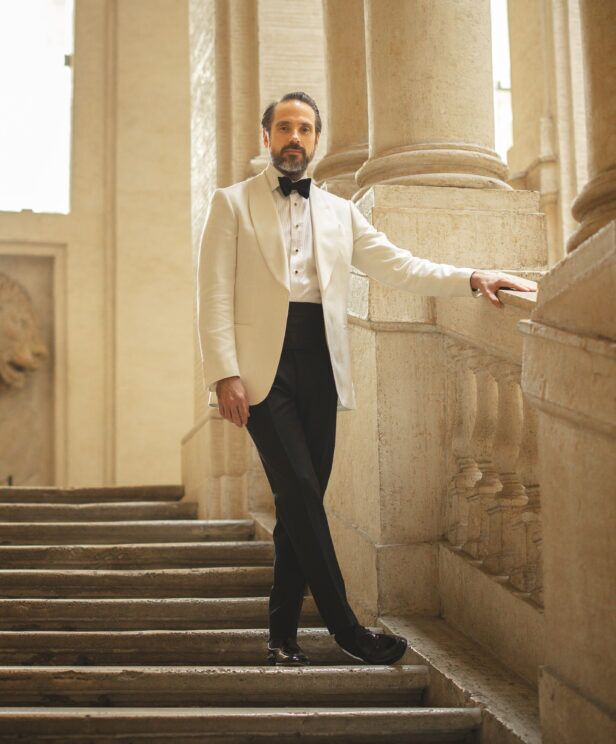 Ivory shawl collar dinner jacket with pleated stud front shirt, black bow tie and cummerbund, worn by a model on stone steps