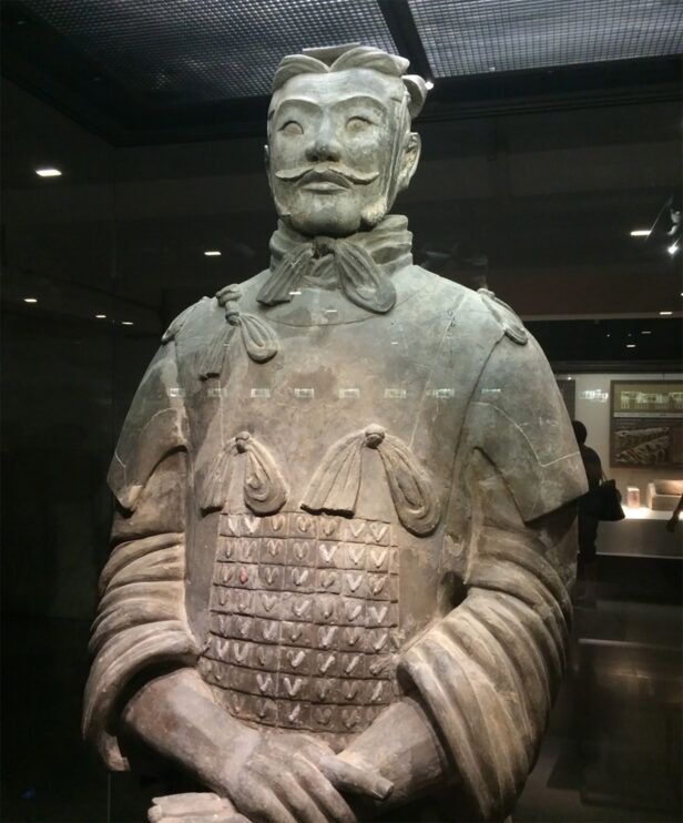 Terracotta warrior statue with knotted neck scarf and scaled armour in a museum case, a styled lifestyle scene