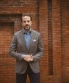 Brown and grey houndstooth wool sport coat with blue chambray shirt, navy trousers and white pocket square, worn by a model