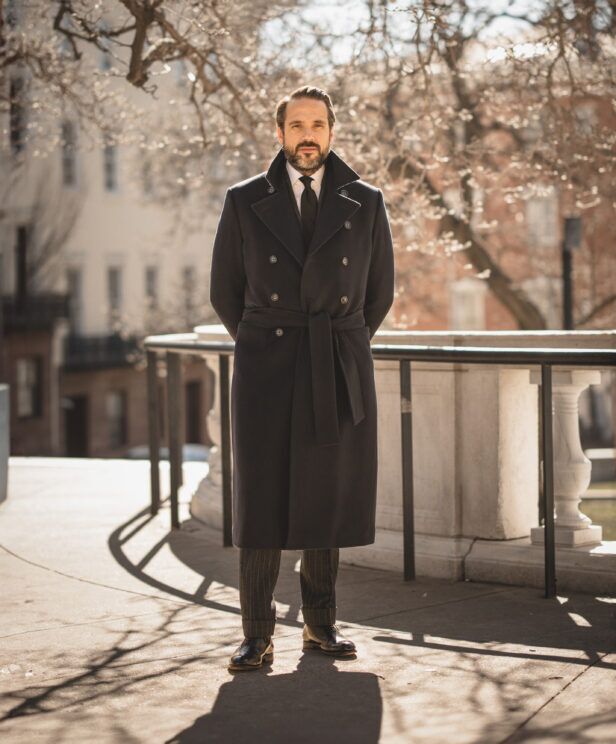Navy double-breasted belted wool greatcoat with striped grey trousers and black shoes, worn by a model outdoors