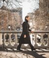 Charcoal double-breasted belted greatcoat with striped trousers and black boots, worn by a model on a city terrace
