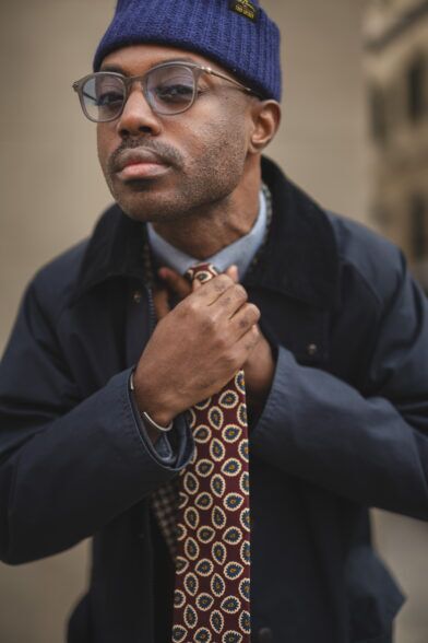 Burgundy silk necktie with blue and cream paisley medallions, worn untied with a navy jacket and beanie by a model