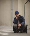 Navy corduroy-collar coat over checked suit, medallion tie and navy beanie, worn by a man crouching on a street