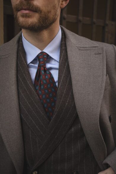 Rust silk necktie with teal medallions, brown chalk stripe waistcoat and taupe overcoat, worn by a model