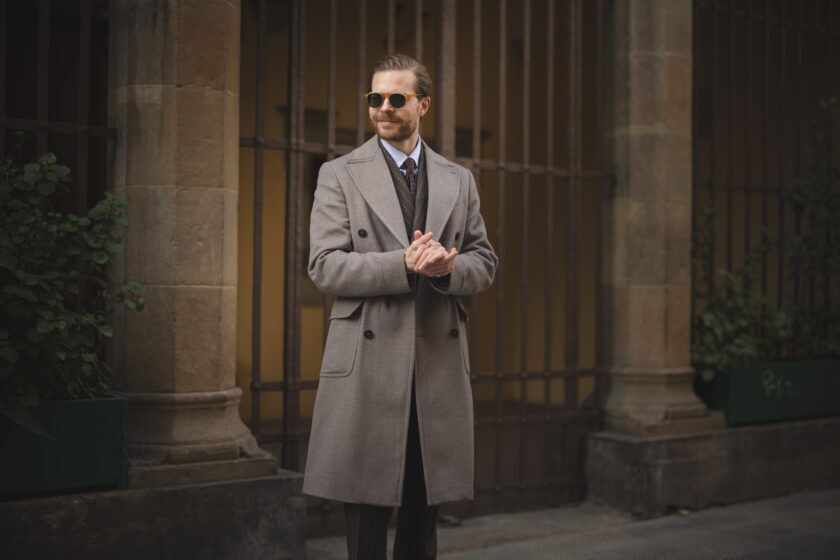 Taupe double-breasted wool overcoat with striped shirt and patterned tie, worn by a man on a stone colonnade street
