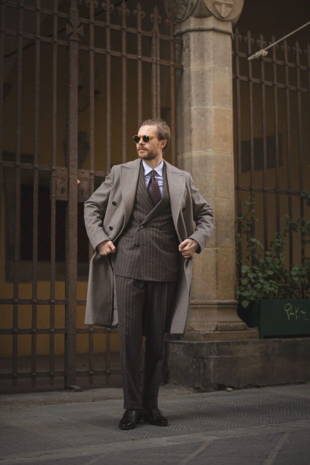 Brown chalk stripe double-breasted suit with taupe overcoat, blue shirt and red medallion tie, worn by a model on a street